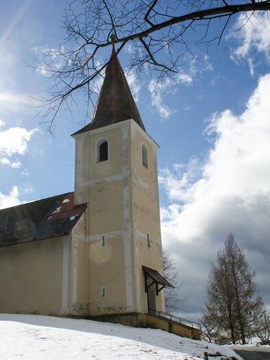 Eine alte Kirche mit einem spitzen Turm steht in einer schneebedeckten Landschaft. Der Himmel ist teils bewölkt, und die Sonne scheint durch die Wolken.