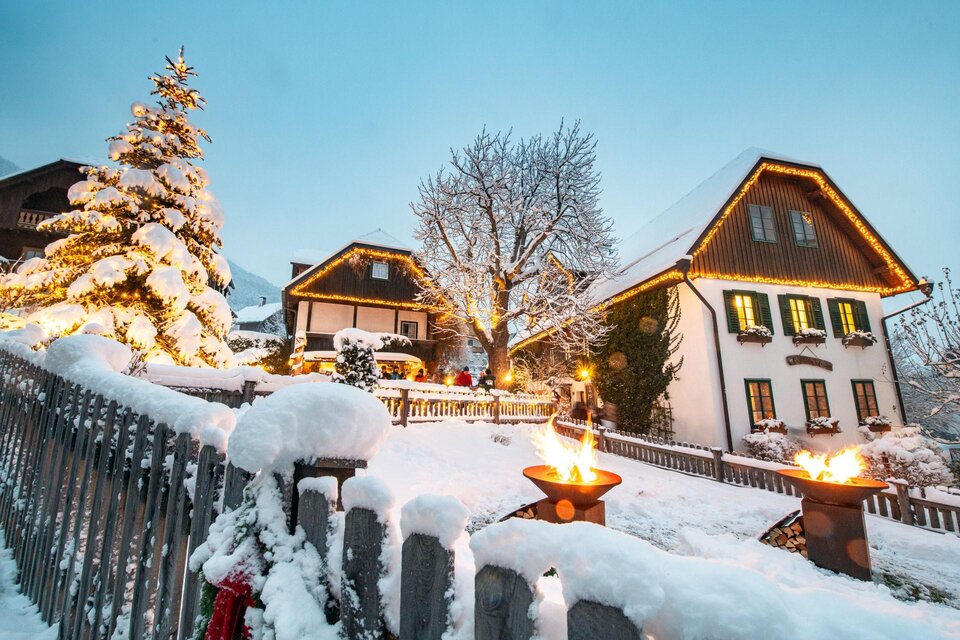 Ein winterliches Dorfhaus mit schneebedecktem Garten. Ein geschmückter Weihnachtsbaum und Fackeln strahlen in der Dämmerung. | © Christoph Huber