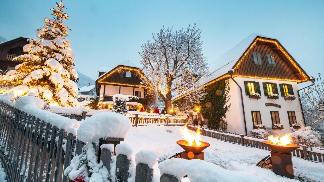 Ein winterliches Dorfhaus mit schneebedecktem Garten. Ein geschmückter Weihnachtsbaum und Fackeln strahlen in der Dämmerung. | © Christoph Huber