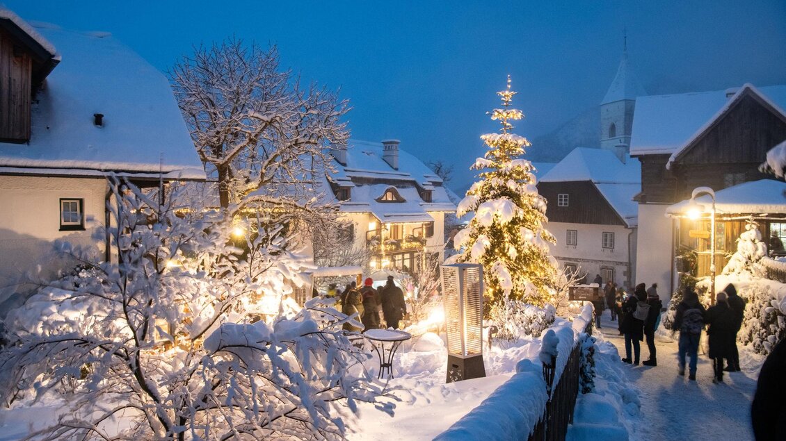 Eine malerische Winterlandschaft mit schneebedeckten Häusern und einem geschmückten Weihnachtsbaum. Menschen schlendern durch die festliche Szenerie unter dem klaren Abendhimmel. | © Christoph Huber