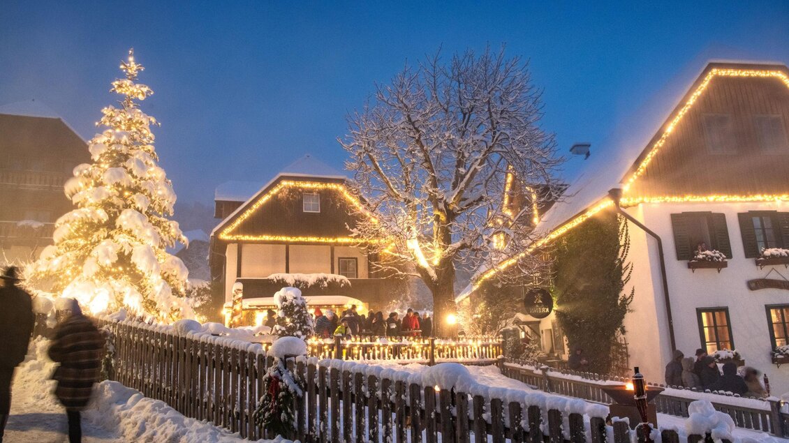 Eine verschneite Winterlandschaft mit festlich beleuchteten Häusern. Ein schön dekorierter Weihnachtsbaum steht in der Mitte, während Menschen vorbeigehen. | © Christoph Huber