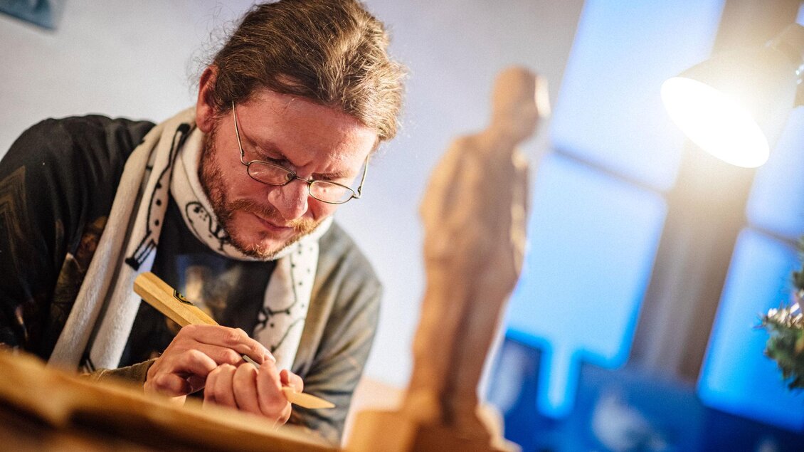 Ein Künstler arbeitet konzentriert an einer Holzskulptur. Im Hintergrund ist eine beleuchtete Figur zu sehen. | © Christoph Huber