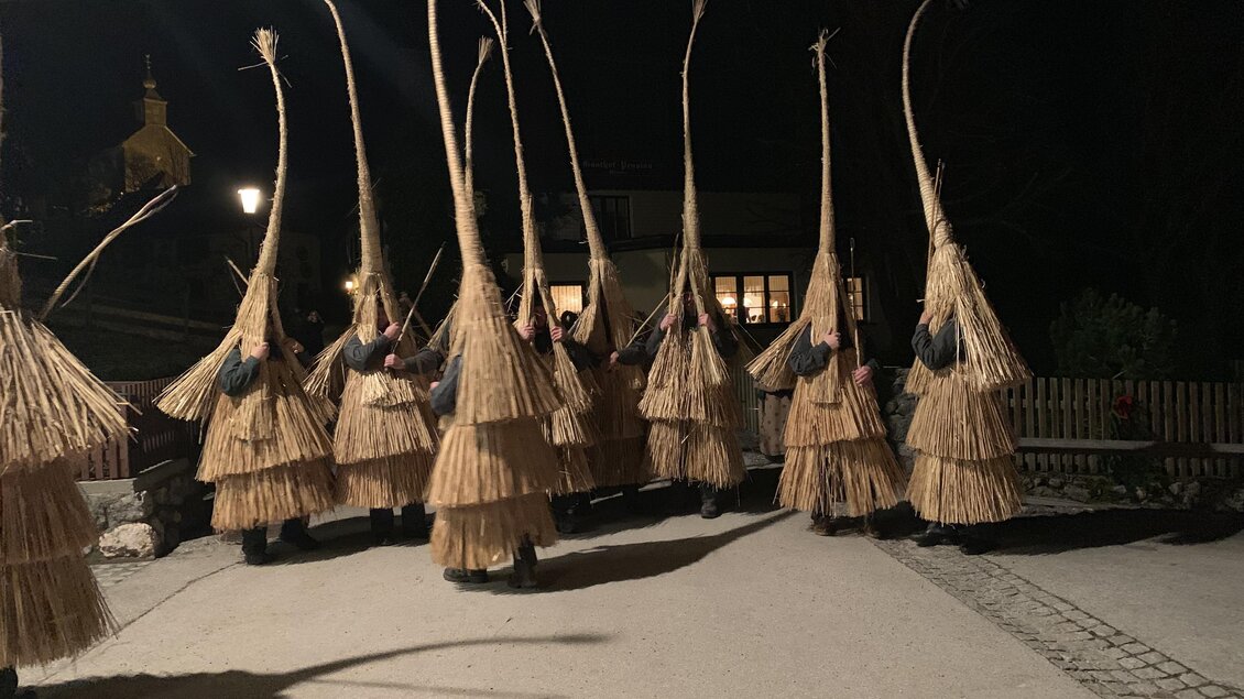 Eine Gruppe von Menschen in traditionellen Kostümen aus Naturmaterialien in einer nächtlichen Umgebung. Sie haben hohe, geflochtene Hüte und stehen in einer Reihe. | © Karlheinz Rainer