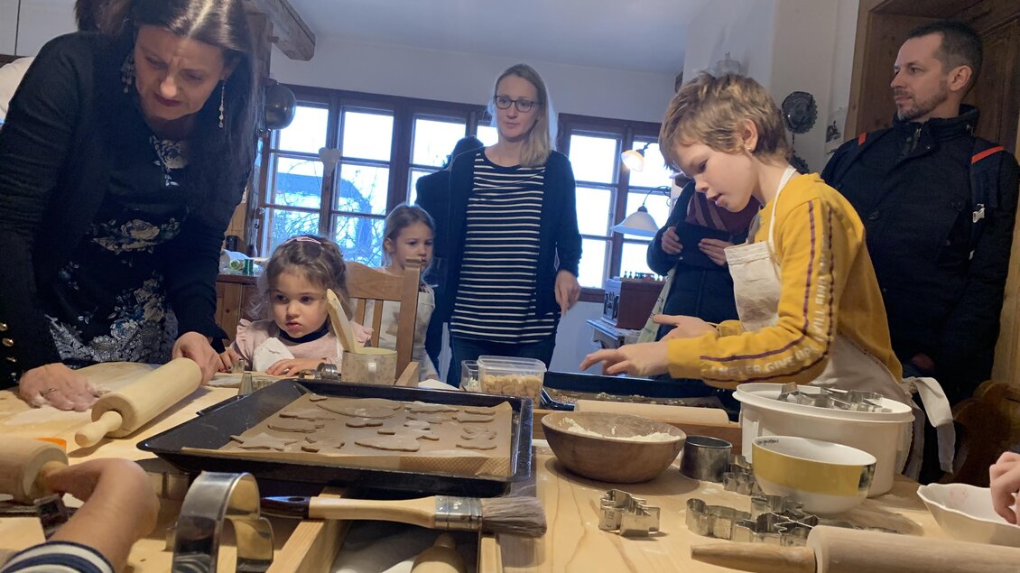 Eine Gruppe von Menschen, darunter Kinder und Erwachsene, versammelt sich in einer Küche zum Plätzchenbacken. Sie arbeiten gemeinsam an einem Tisch mit Teig, Förmchen und Backblechen. | © Karlheinz Rainer
