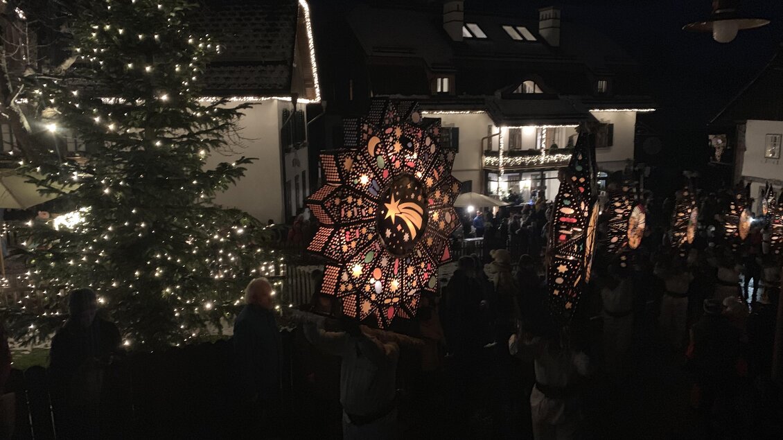 Eine festliche Szene mit leuchtenden Laternen und einem Weihnachtsbaum. Menschen stehen in einer Gruppe und feiern in der Dunkelheit. | © Karlheinz Rainer
