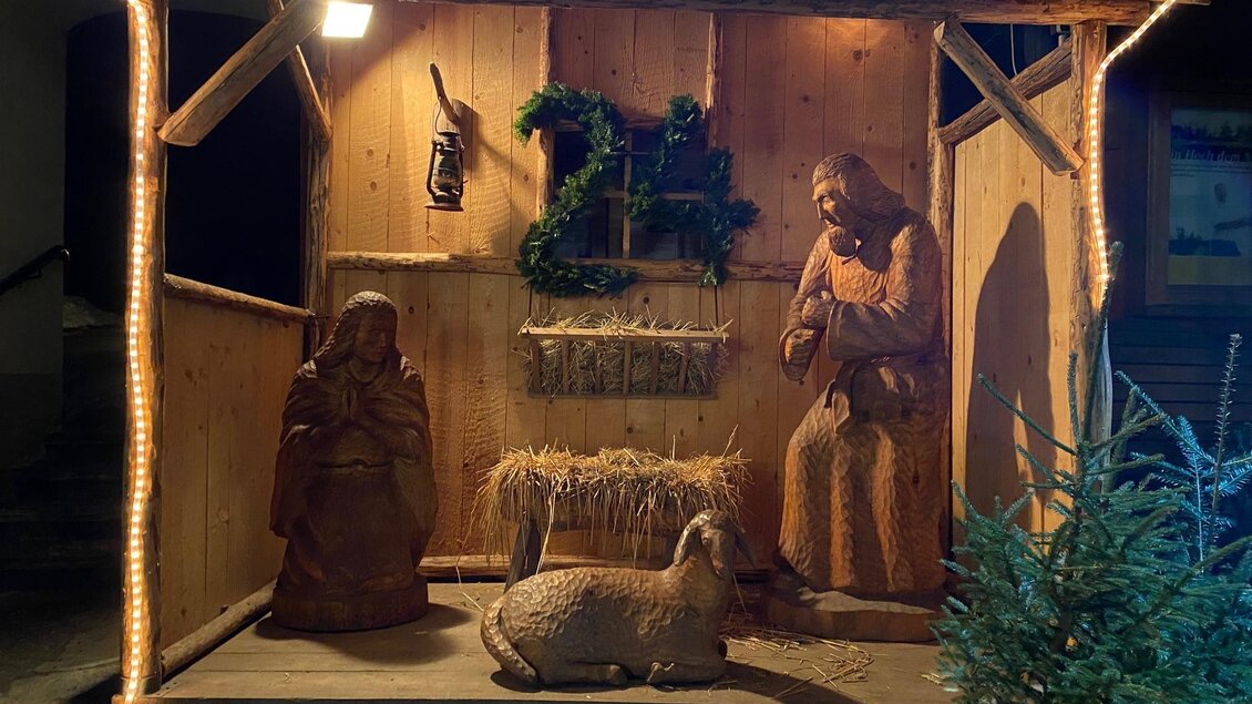 Eine weihnachtliche Krippe aus Holz mit Figuren von Maria, Josef und einem Schaf. Die Szene ist festlich beleuchtet und mit Tannenzweigen geschmückt. | © Alexandra Wippel
