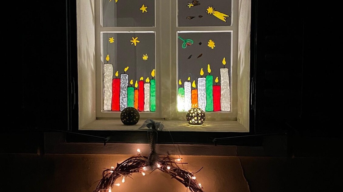 Ein Fenster mit bunten Weihnachtsdekorationen, darunter Kerzen und Sterne. Ein Lichterkranz schmückt den unteren Teil des Fensters. | © Alexandra Wippel