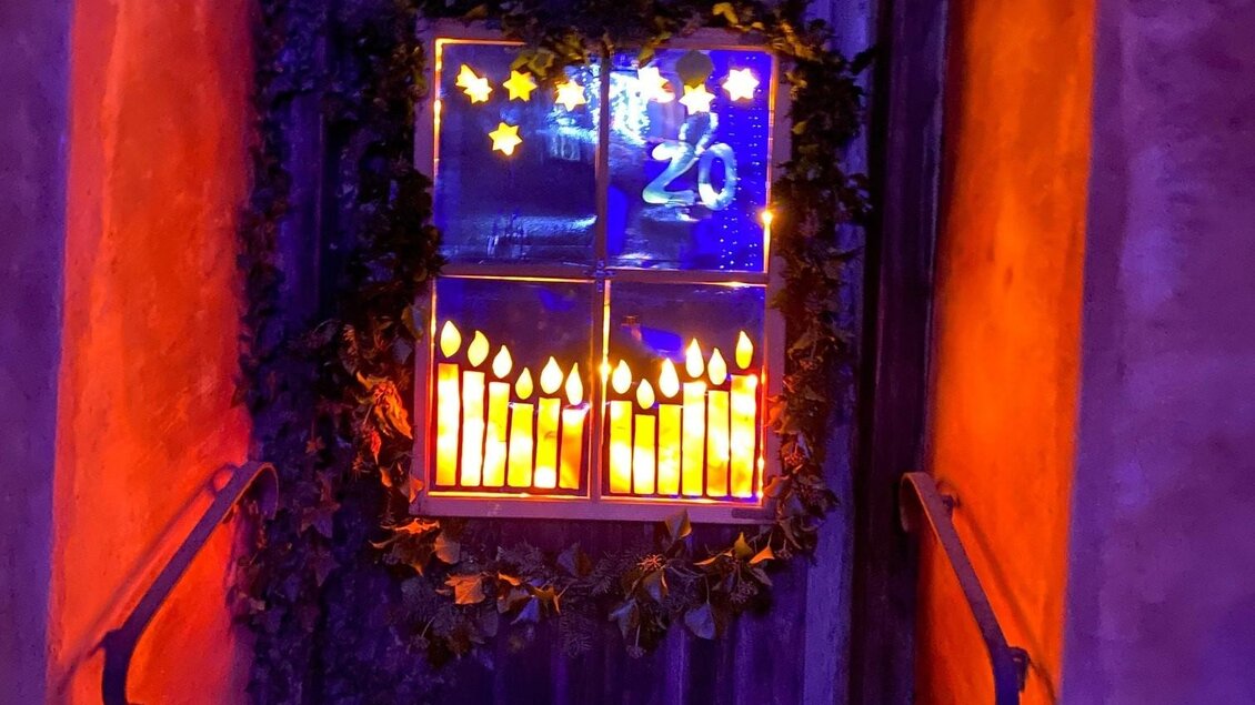 Ein beleuchtetes Fenster mit festlicher Dekoration und Kerzen auf einer Mauer, umgeben von grünem Efeu. Der Eingang wird in warmen, leuchtenden Farben hervorgehoben. | © Alexandra Wippel