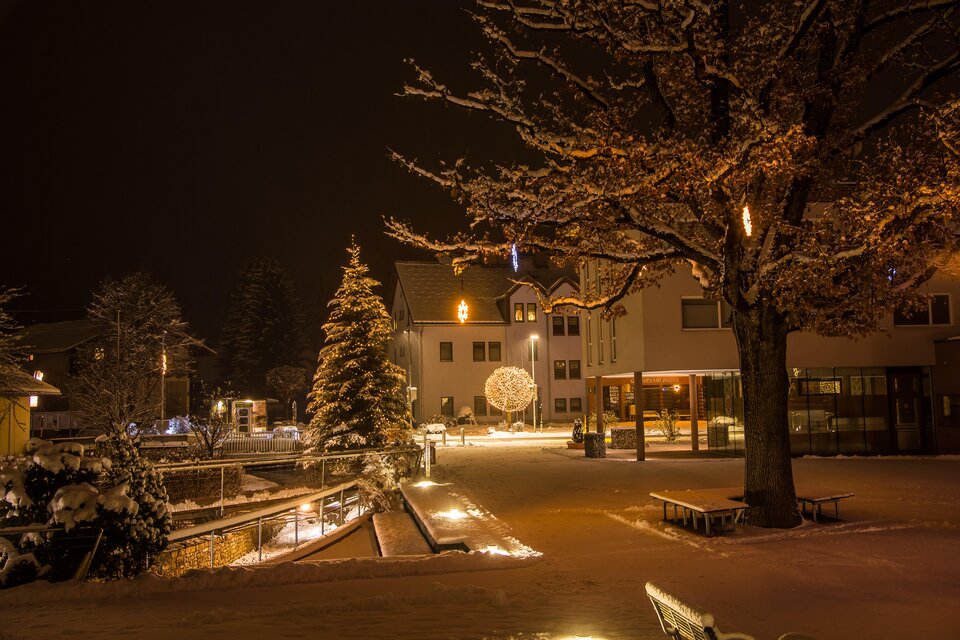 Ein verschneiter Platz bei Nacht mit festlicher Beleuchtung. Im Hintergrund steht ein geschmückter Weihnachtsbaum und die umliegenden Gebäude sind sanft erleuchtet. | © Gemeinde Aigen im Ennstal
