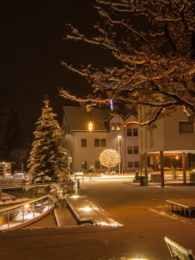 Ein verschneiter Platz bei Nacht mit festlicher Beleuchtung. Im Hintergrund steht ein geschmückter Weihnachtsbaum und die umliegenden Gebäude sind sanft erleuchtet. | © Gemeinde Aigen im Ennstal
