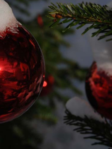 Rote Weihnachtsbaumkugeln, die mit Schnee bedeckt sind, hängen an einer Tannenzweigen. Im Hintergrund sind Schatten und Lichtspiele sichtbar. | © Pirker