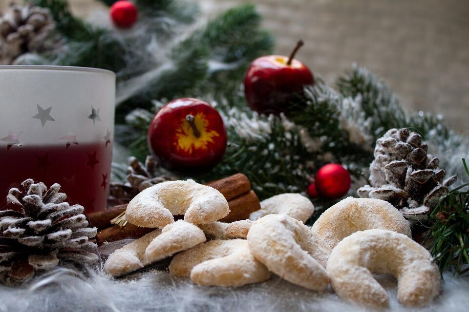 Eine winterliche Dekoration mit leckeren Plätzchen, Tannenzapfen und Äpfeln. Im Hintergrund befindet sich ein Glas mit einer weihnachtlichen Getränkespezialität. | © Pixabay