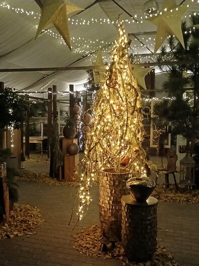Eine festlich dekorierte Umgebung mit glitzernden Lichtern und einem dekorativen Baum. Der Boden ist mit goldenen Blättern bedeckt und die Stimmung ist warm und einladend. | © Gärtnerei Steffan