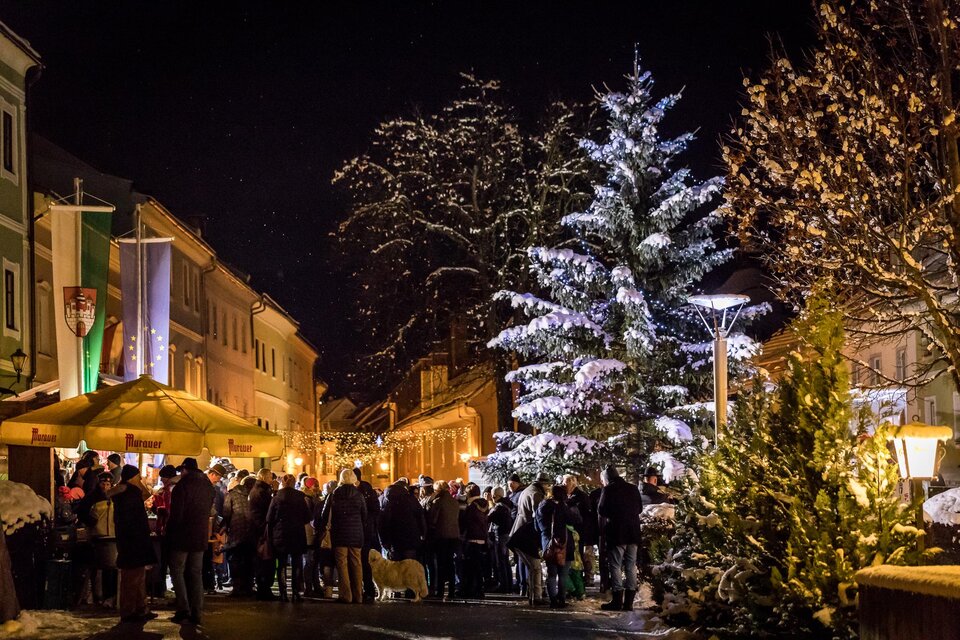 Ein festlicher Weihnachtsmarkt bei Nacht mit vielen Menschen, die sich versammelt haben. Im Vordergrund steht ein schön beleuchteter Weihnachtsbaum.