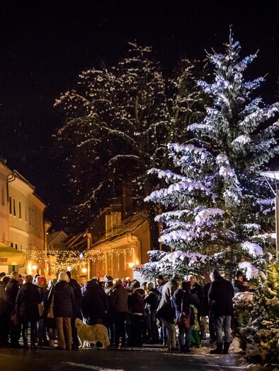 Ein festlicher Weihnachtsmarkt bei Nacht mit vielen Menschen, die sich versammelt haben. Im Vordergrund steht ein schön beleuchteter Weihnachtsbaum.
