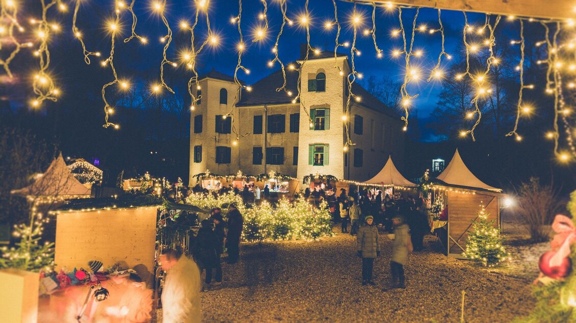 Ein festlicher Weihnachtsmarkt mit hellen Lichtern und vielen Menschen. Im Hintergrund ist ein beleuchtetes Gebäude und Zelte zu sehen. | © Harald Tauderer Photography
