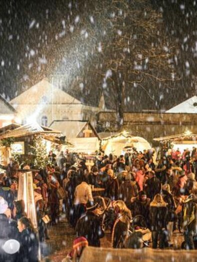 Ein festlicher Weihnachtsmarkt bei Nacht, beleuchtet mit Lichtern und umgeben von Schnee. Menschen genießen die festliche Atmosphäre und die Stände mit verschiedenen Angeboten.