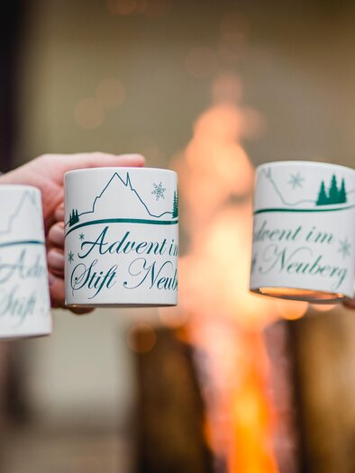 Drei Personen halten Tassen mit festlichen Motiven in der Nähe eines Lagerfeuers. Die Tassen zeigen die Aufschriften „Advent im Stift Neutal“ und „Advent in Tirol“. | © nicoleseiser.at