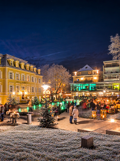 Eine festlich beleuchtete Szene mit einem prächtigen gelben Gebäude im Hintergrund. Im Vordergrund stehen Weihnachtsstände und festlich geschmückte Bäume im Schnee. | © BG Kultur und Entwicklungs GmbH
