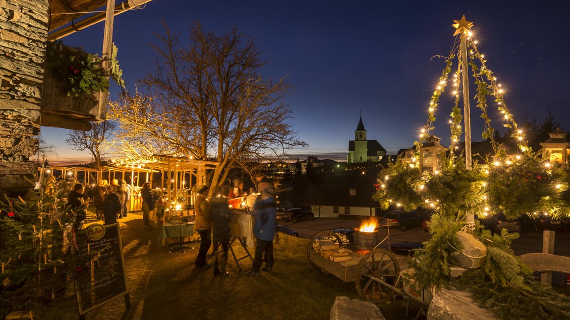 Ein festlich geschmückter Platz bei Dämmerung mit Lichtern und einem Weihnachtsbaum. Menschen stehen zusammen und genießen die stimmungsvolle Atmosphäre. | © Oststeiermark Tourismus