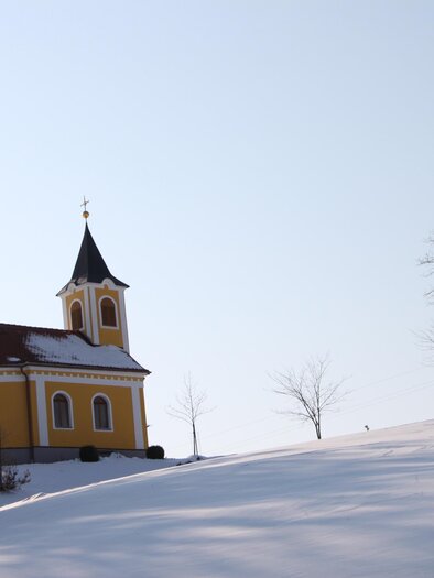 The Mother-Anna Chapel amidst the snowy landscape in Haselbach near Unterlamm. The warm yellow tone of the facade creates a beautiful contrast to the white snow and gives the quiet winter scene a particularly peaceful atmosphere. | © Kapellengemeinschaft Haselbach