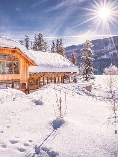 Ein wunderschönes Holzhaus in einer schneebedeckten Landschaft. Die Sonne strahlt über die Berge und schafft eine malerische Winterkulisse. | © Kohlröserlhütte_Genuss am See