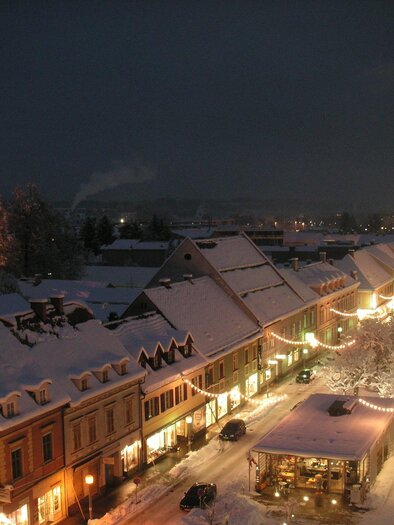 A picturesque town at night, covered in snow and festive lighting. In the background stands a tall tower and the street is full of lights. | © FantaFilm