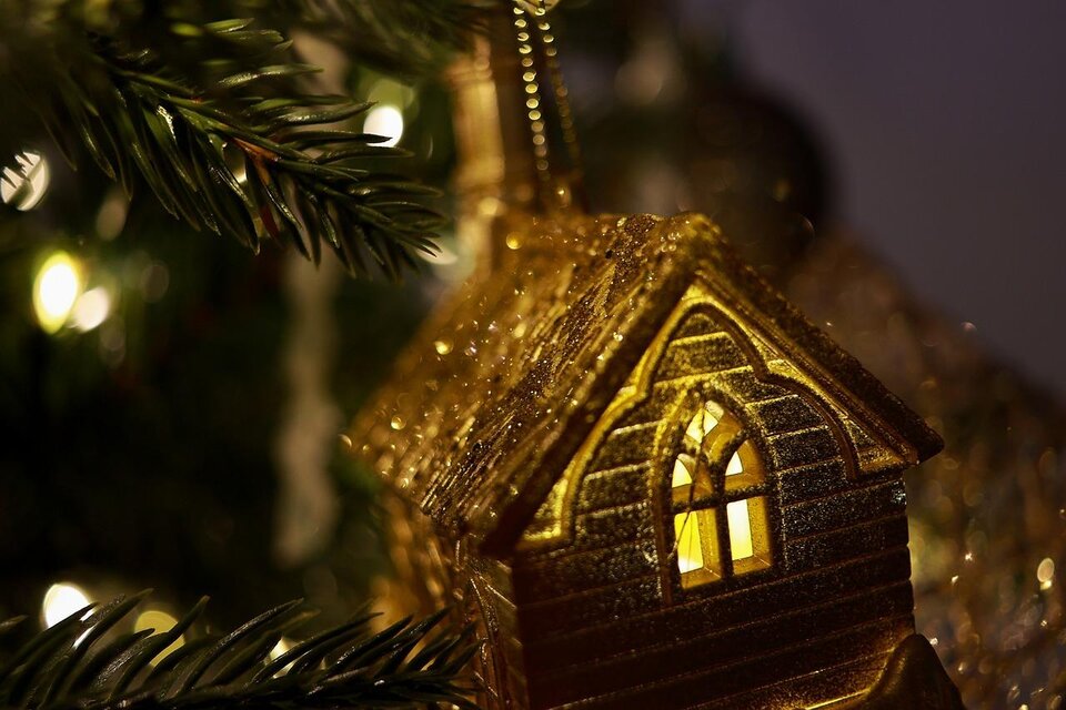 Ein goldenes Weihnachtsbaumornament in Form eines Hauses mit leuchtendem Fenster. Im Hintergrund schimmern dekorative Lichter. | © Pixabay.com