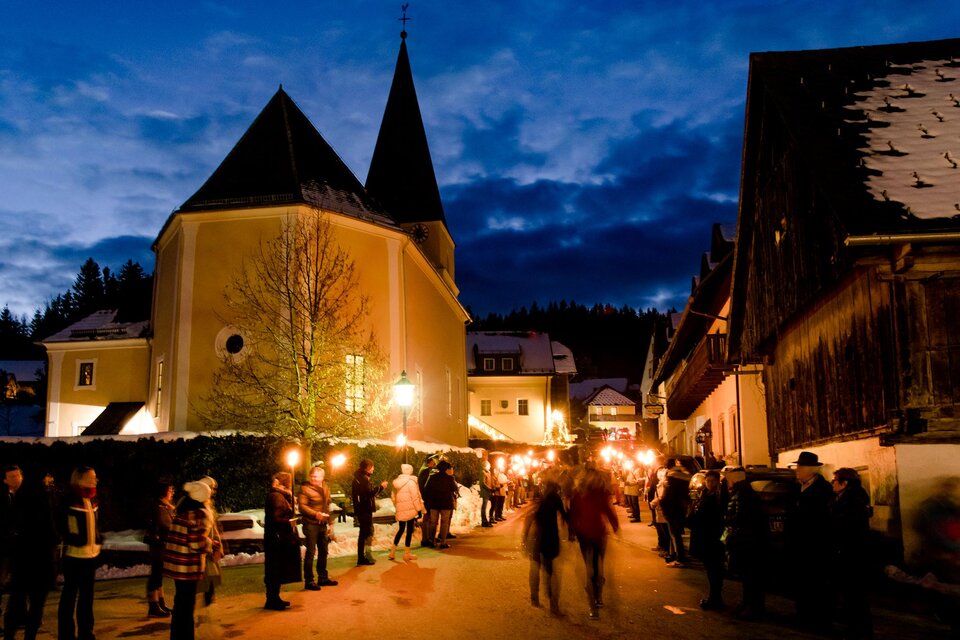 Eine festliche Szene in einem winterlichen Dorf mit einer Kirche im Hintergrund. Menschen stehen mit Fackeln auf der Straße unter einem klaren Abendhimmel. | © Region Graz-Mias Photoart
