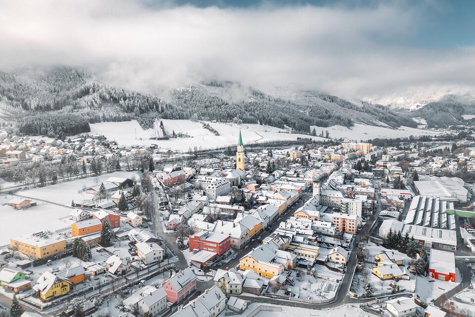 Eine verschneite Stadt mit bunten Häusern und einem Kirchturm in der Mitte. Die umliegenden Berge sind von Wolken umhüllt.