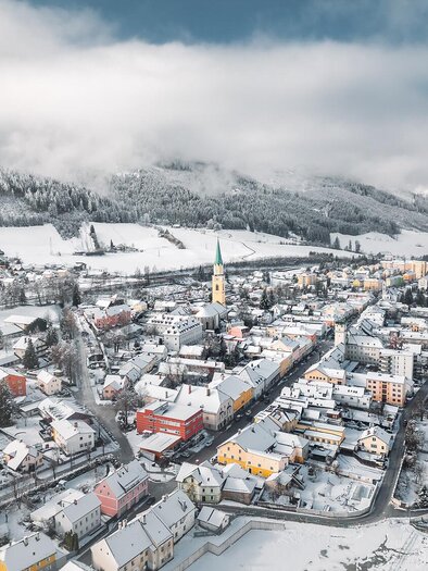 Eine verschneite Stadt mit bunten Häusern und einem Kirchturm in der Mitte. Die umliegenden Berge sind von Wolken umhüllt.