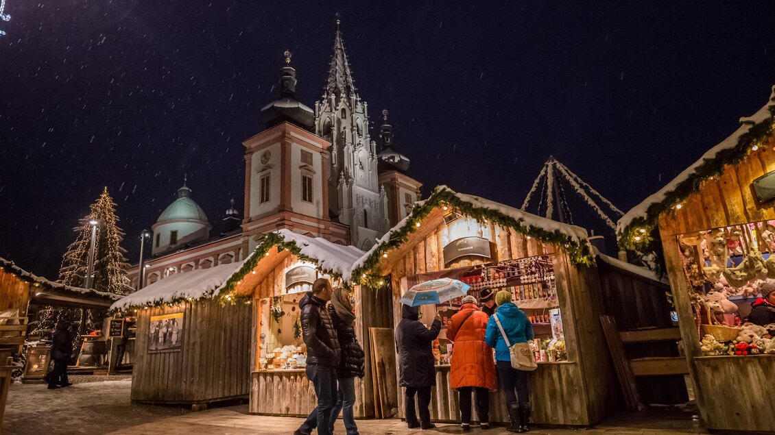 Ein festlicher Weihnachtsmarkt bei Nacht mit beleuchteten Holzhütten. Im Hintergrund ist eine beeindruckende Kirche zu sehen, während Besucher die Stände erkunden. | © Fred Lindmoser