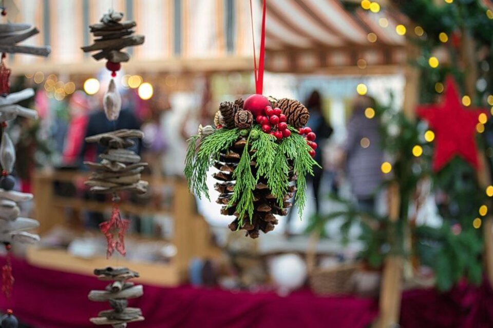 Ein dekorativer Tannenzapfen mit grünem Blattwerk und roten Beeren hängt in einem festlichen Umfeld. Im Hintergrund sind verschwommene Lichter und Stände eines Weihnachtsmarkts zu sehen. | © Pixabay