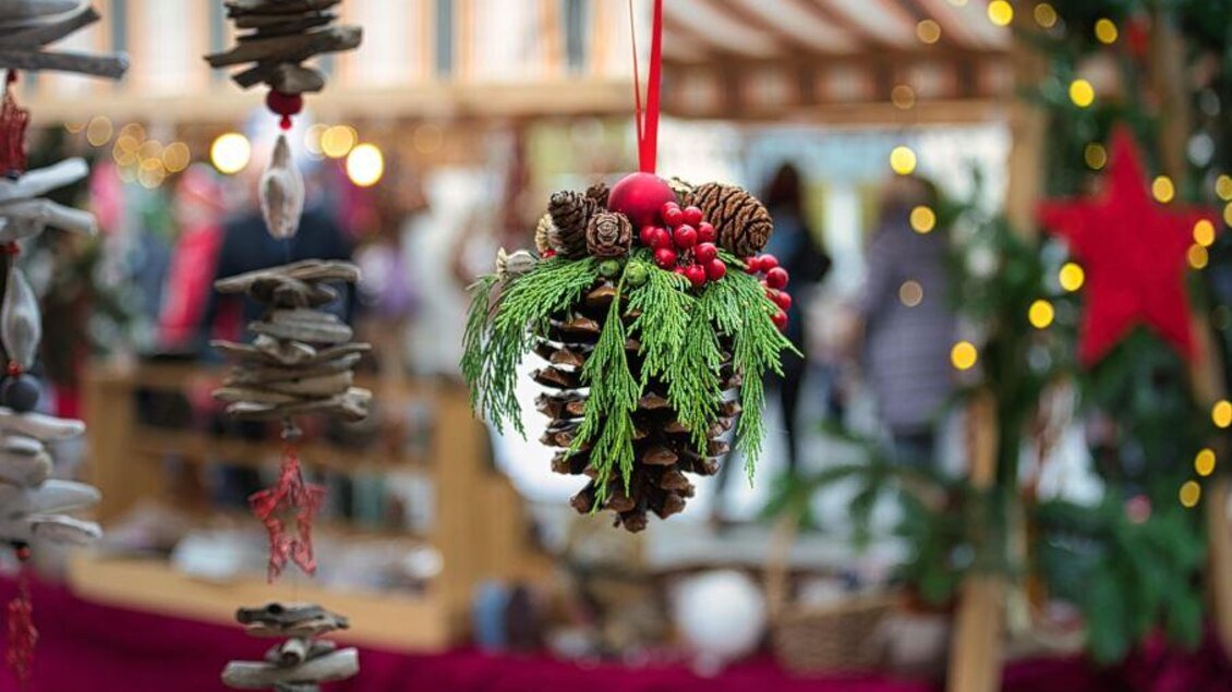 Ein dekorativer Tannenzapfen mit grünem Blattwerk und roten Beeren hängt in einem festlichen Umfeld. Im Hintergrund sind verschwommene Lichter und Stände eines Weihnachtsmarkts zu sehen. | © Pixabay
