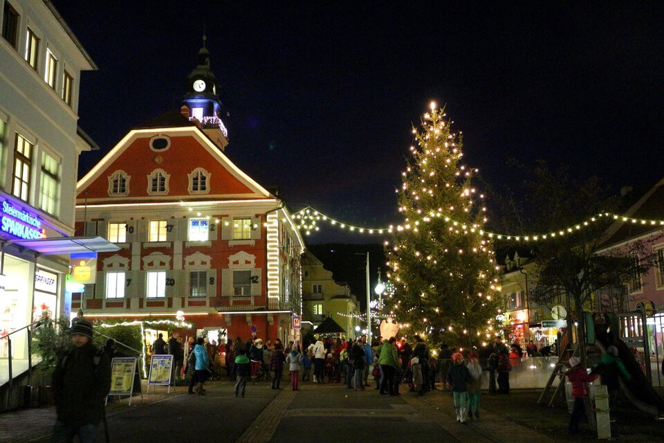 Eine festliche Stadtansicht bei Nacht mit einem beleuchteten Weihnachtsbaum. Es gibt viele Menschen auf der Straße, die die festliche Atmosphäre genießen. | © Gemeinde DL