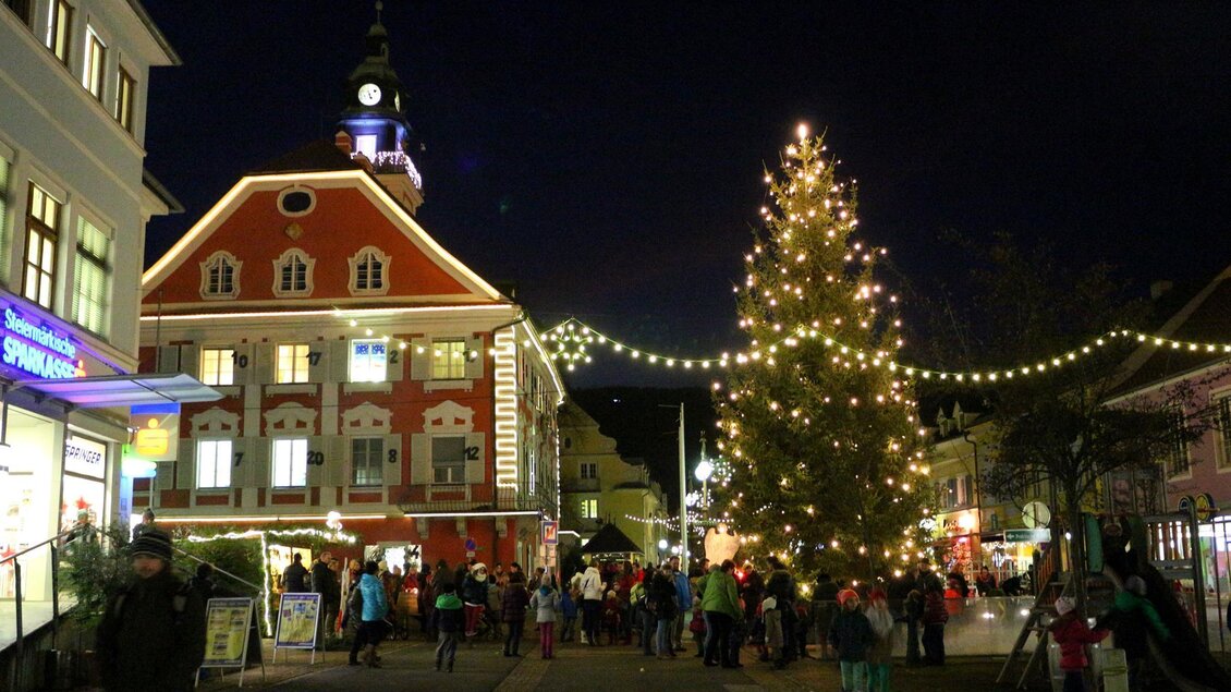 Eine festliche Stadtansicht bei Nacht mit einem beleuchteten Weihnachtsbaum. Es gibt viele Menschen auf der Straße, die die festliche Atmosphäre genießen. | © Gemeinde DL