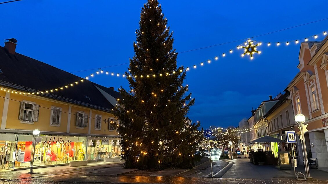 Ein festlich geschmückter Weihnachtsbaum steht in der Stadtmitte. Die Straßen sind mit Lichtern dekoriert und der Himmel ist dunkelblau.