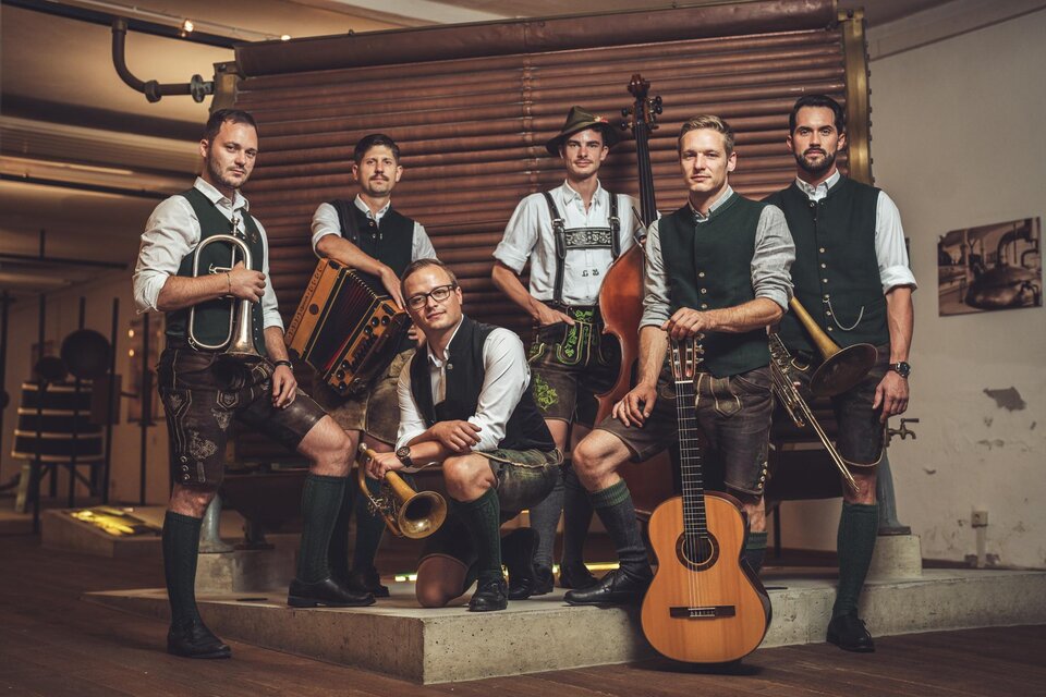 Eine Gruppe von sechs Männern in traditioneller bayerischer Tracht steht zusammen. Sie halten Musikinstrumente und posieren in einem rustikalen Innenraum. | © Reithofer Media