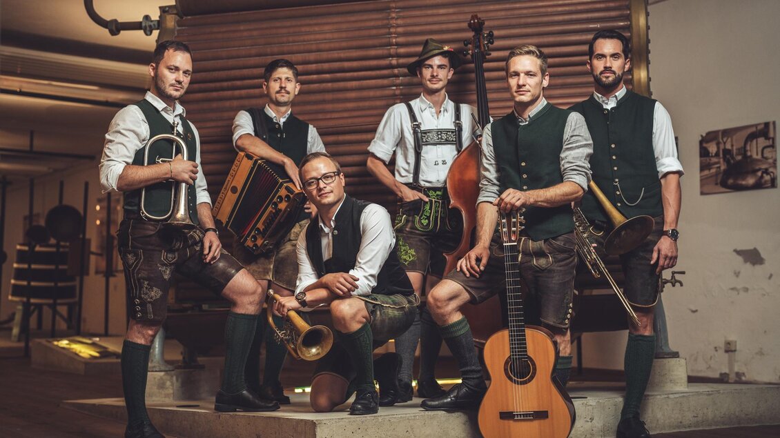 Eine Gruppe von sechs Männern in traditioneller bayerischer Tracht steht zusammen. Sie halten Musikinstrumente und posieren in einem rustikalen Innenraum. | © Reithofer Media