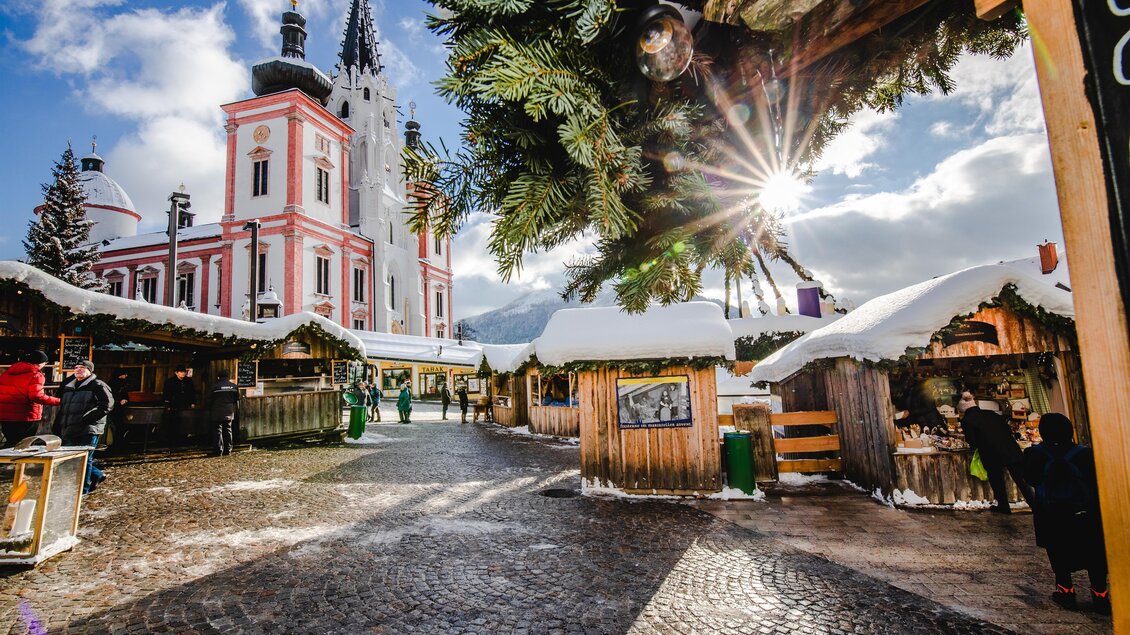 Ein malerischer Weihnachtsmarkt mit belebten Holzständen und festlicher Dekoration. Im Hintergrund ist eine beeindruckende pinke Kirche unter einem klaren Himmel zu sehen. | © Fred Lindmoser