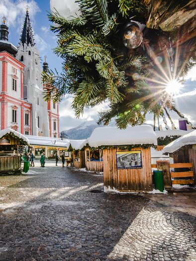 A picturesque Christmas market with bustling wooden stalls and festive decorations. In the background, there is an impressive pink church under a clear sky. | © Fred Lindmoser
