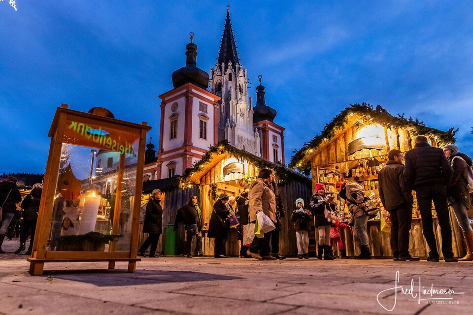 Ein festlicher Markt mit beleuchteten Ständen und vielen Besuchern. Im Hintergrund ist eine beeindruckende Kirche im Abendlicht zu sehen. | © Fred Lindmoser
