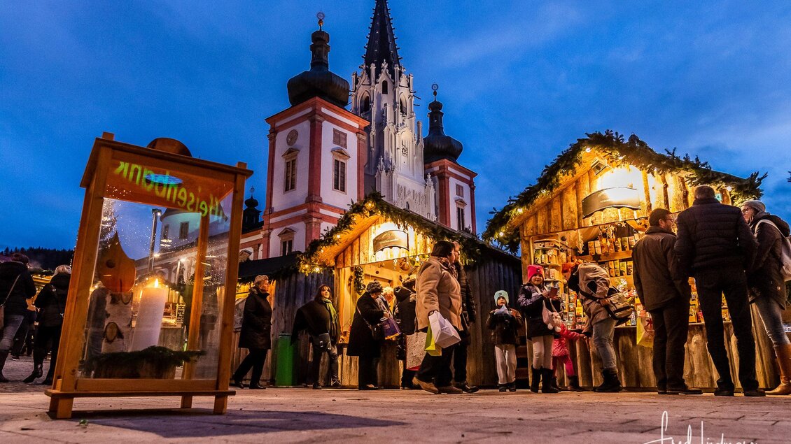 Ein festlicher Markt mit beleuchteten Ständen und vielen Besuchern. Im Hintergrund ist eine beeindruckende Kirche im Abendlicht zu sehen. | © Fred Lindmoser