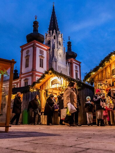 Ein festlicher Markt mit beleuchteten Ständen und vielen Besuchern. Im Hintergrund ist eine beeindruckende Kirche im Abendlicht zu sehen. | © Fred Lindmoser