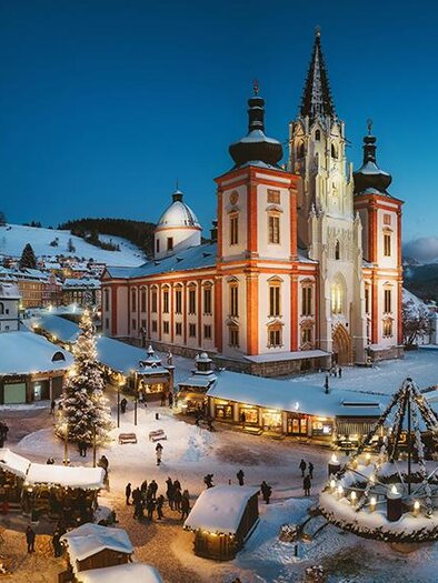 A choir of men in long, dark robes sings in a church. In the background, a Christmas tree and elaborate architecture can be seen. | © Fred Lindmoser