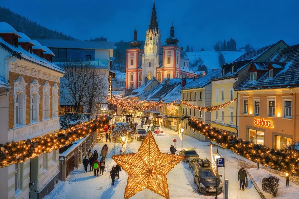 Eine festlich dekorierte Straße in einer winterlichen Stadt mit einem großen leuchtenden Stern. Im Hintergrund ist eine Kirche und die Umgebung ist mit Schnee bedeckt. | © Fred Lindmoser