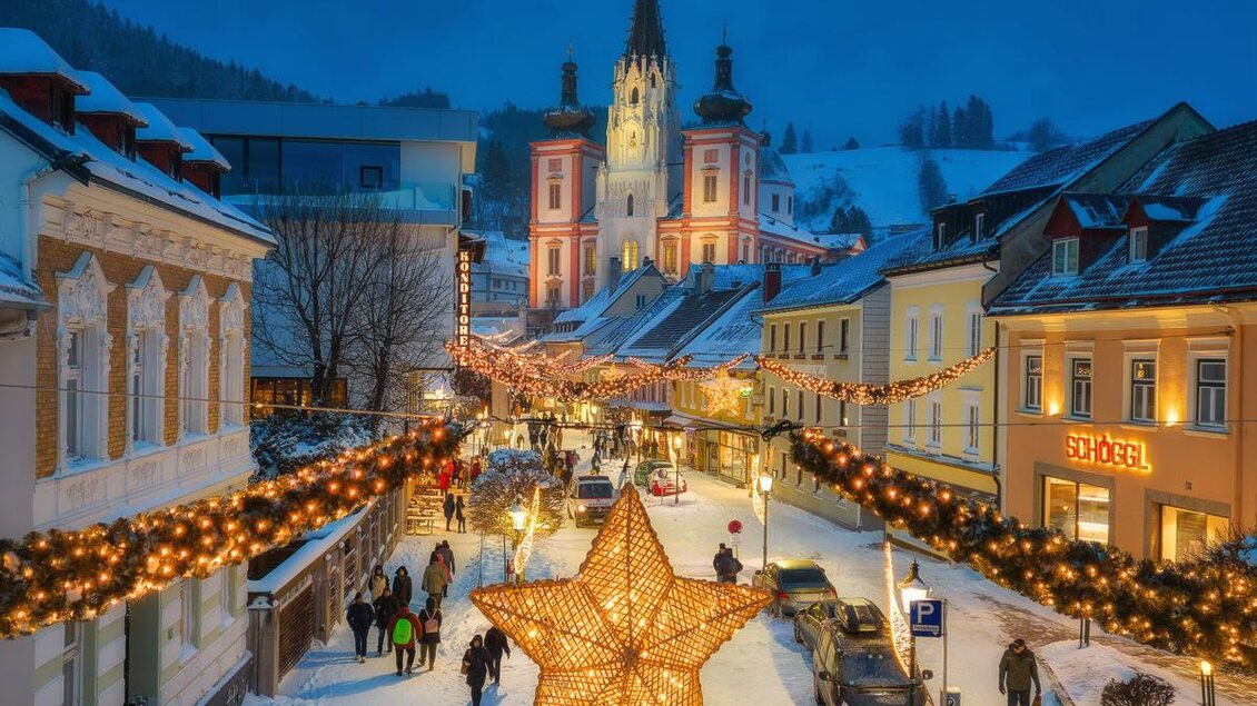 Eine festlich dekorierte Straße in einer winterlichen Stadt mit einem großen leuchtenden Stern. Im Hintergrund ist eine Kirche und die Umgebung ist mit Schnee bedeckt. | © Fred Lindmoser