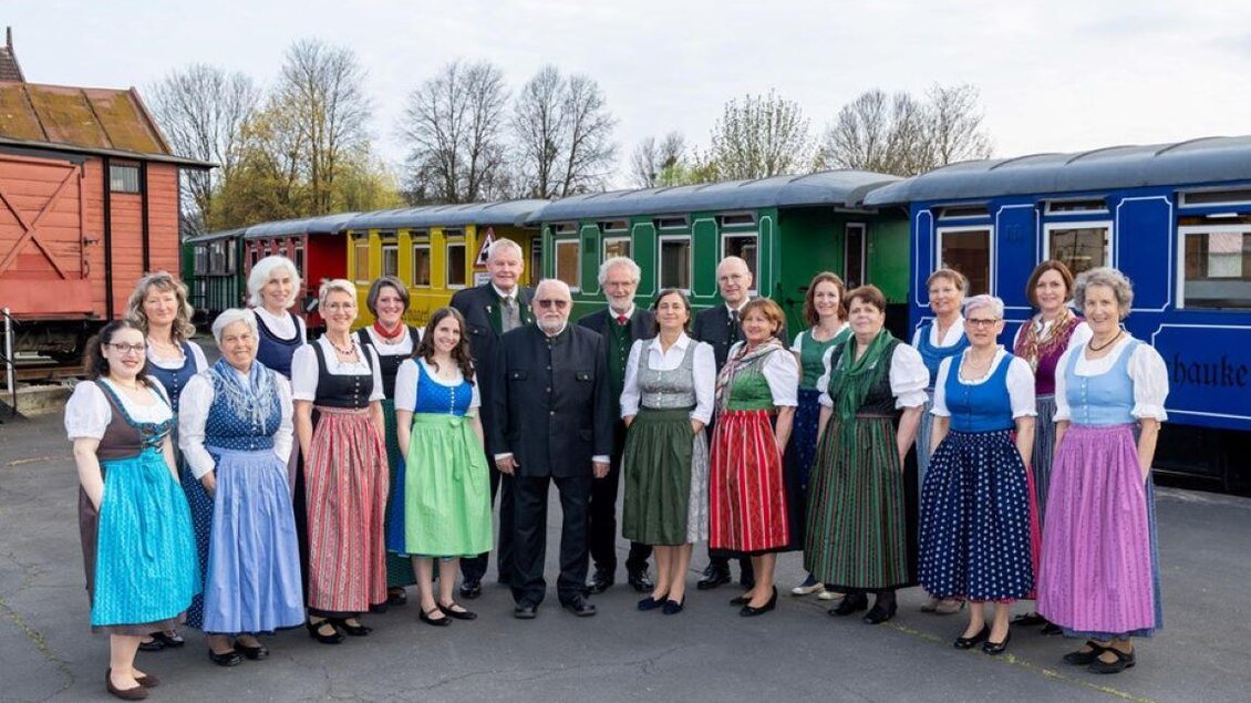 Eine Gruppe von Menschen in traditionellen Trachten steht vor bunten Zugwaggons. Die Atmosphäre ist festlich und freundlich. | © StainZeit - Stainzer Chor