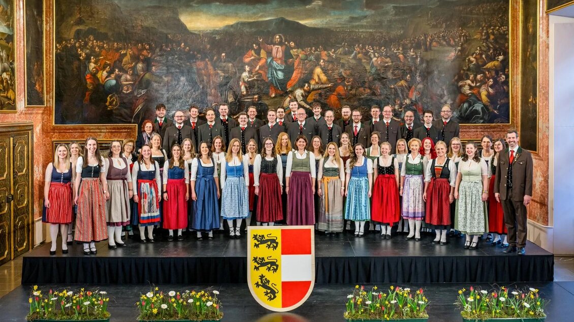 Eine Gruppe von Sängerinnen und Sängern steht auf einer Bühne, gekleidet in traditionelle Trachten. Im Hintergrund ist ein großes Gemälde und ein Wappen zu sehen. | © StainZeit - Grazer Chor