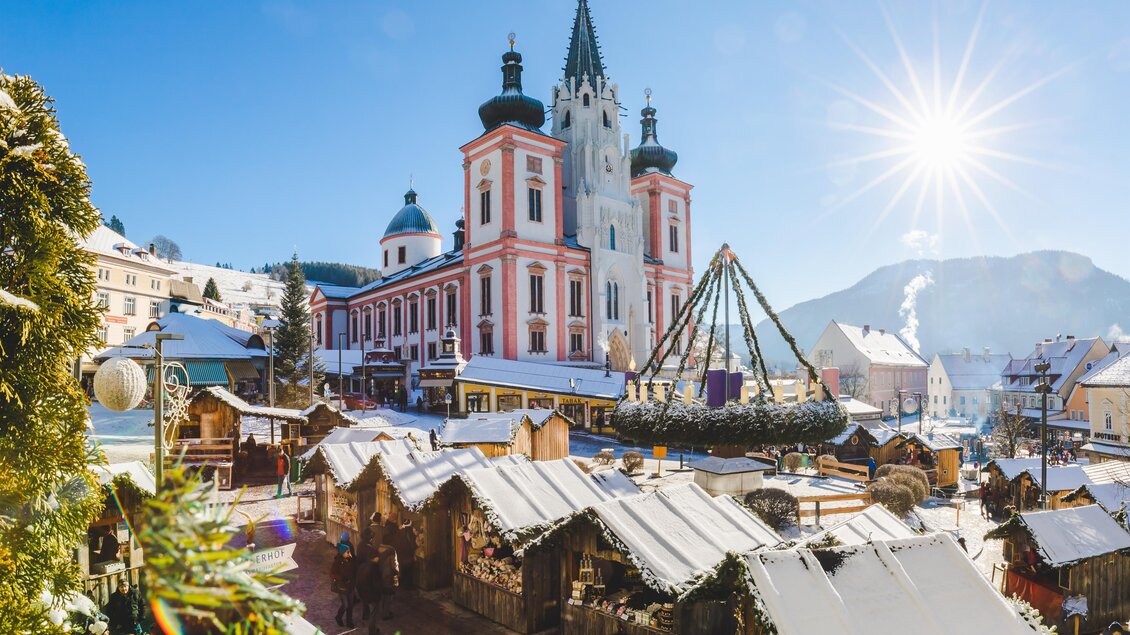 Ein malerischer Weihnachtsmarkt mit Holzständen und schneebedeckten Dächern. Im Hintergrund strahlt ein wunderschönes Gebäude unter klarem Himmel und Sonnenschein. | © Fred Lindmoser